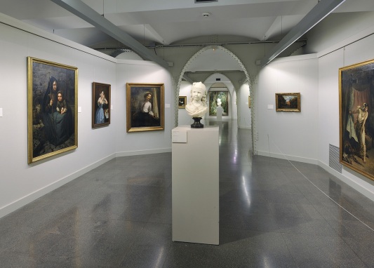 Sala de exposición de pinturas, con un busto en el centro y con una puerta a otra sala
